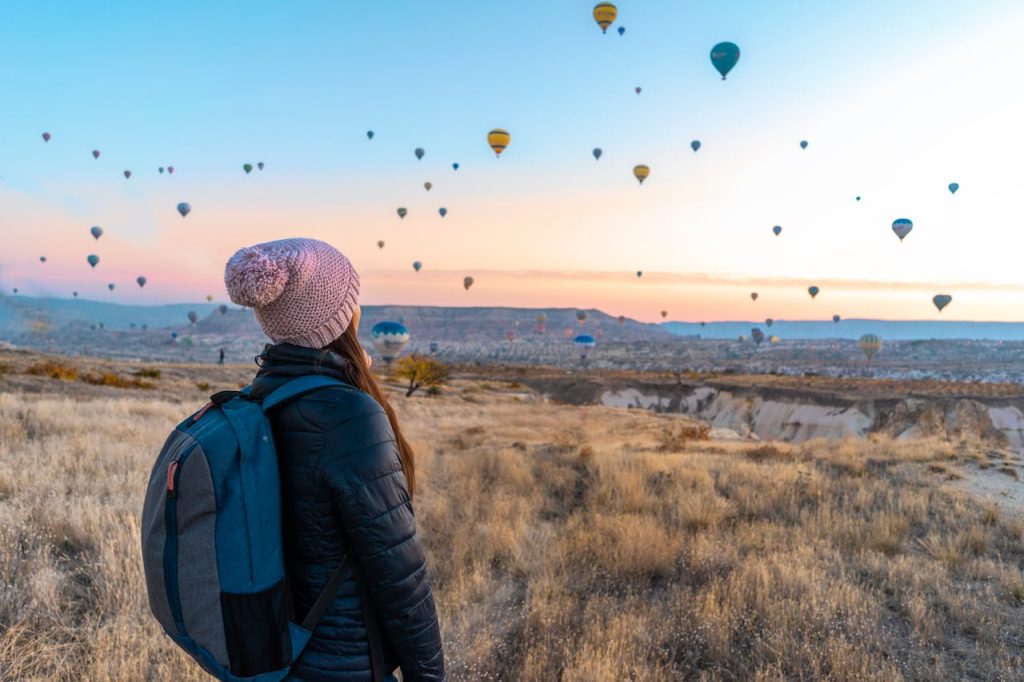 A woman with a backpack standing there while watching a high number of air balloons flying.