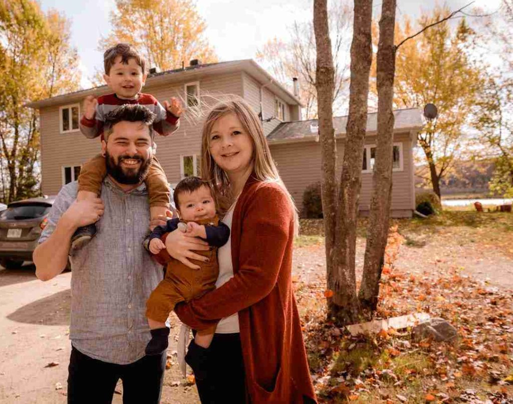 A mum and a dad holding their two kids while standing in front of their house, posing for the camera.