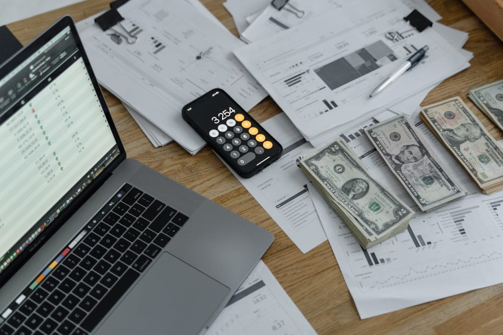 A table with a laptop and an open calculation, a phone used as calculator, many finance documents and money on it.