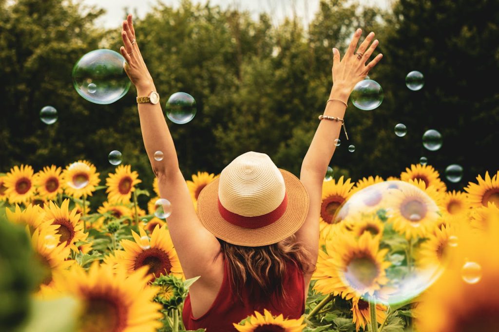 A happy woman, who is standing in a sunflower field holding her arms up and being surrounded by soap bubbles.