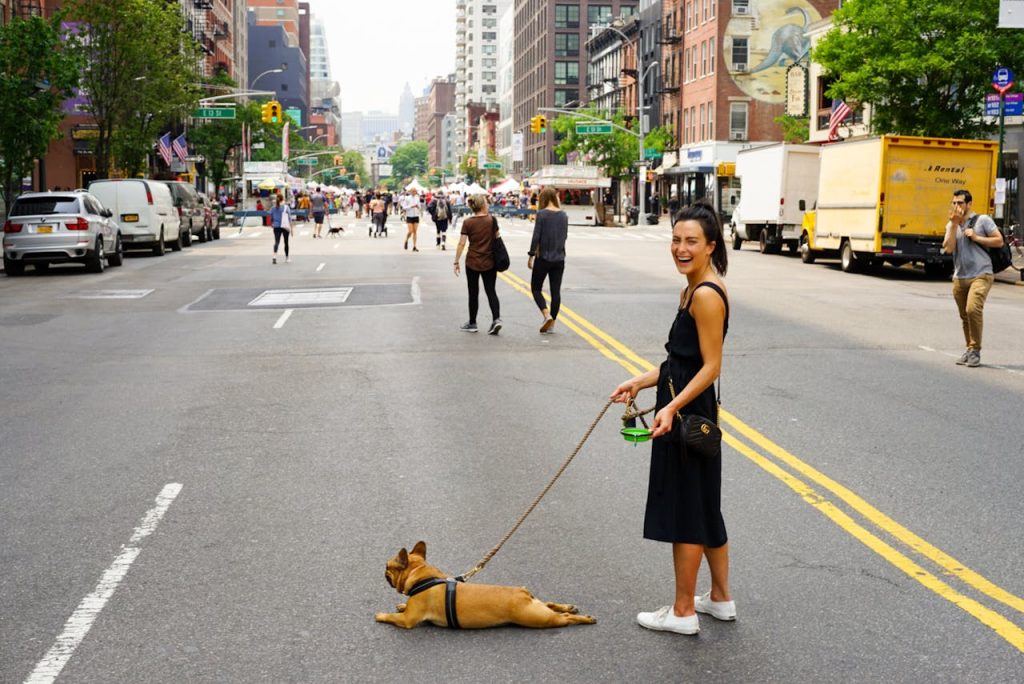 A laughing woman is standing on the middle of the streets of New York while her dog lies right on that street, ready to be served the water, she is holding in her hand.