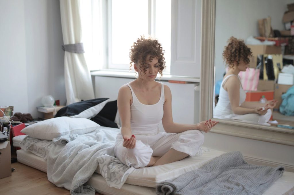 Woman sitting on her bed in her messy room, where you can see a lot of stuff piled up due to the mirror.