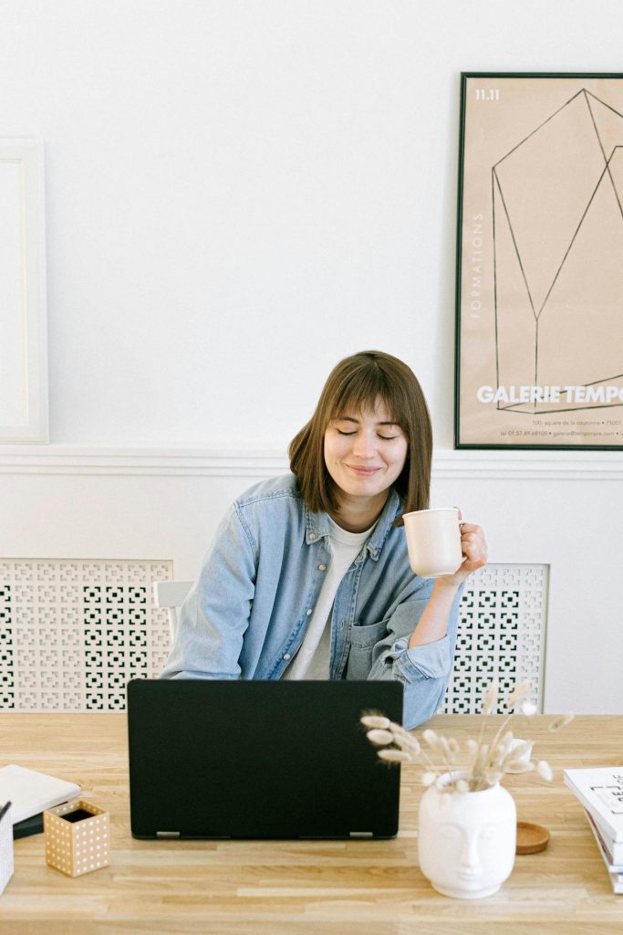 A woman working on her laptop, holding a coffee in her hand and smiling